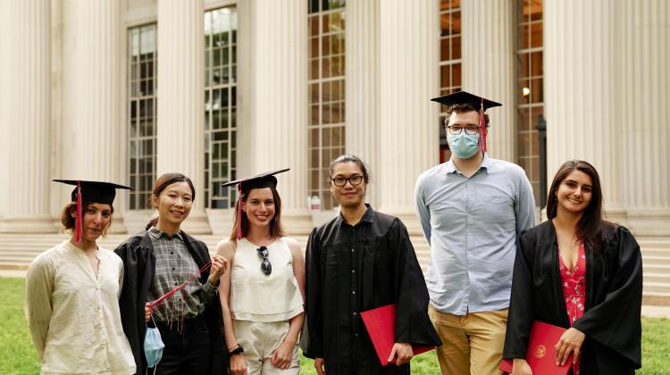 Five graduates standing in front of Building 10, some wearing graduate caps, pose in a row. 